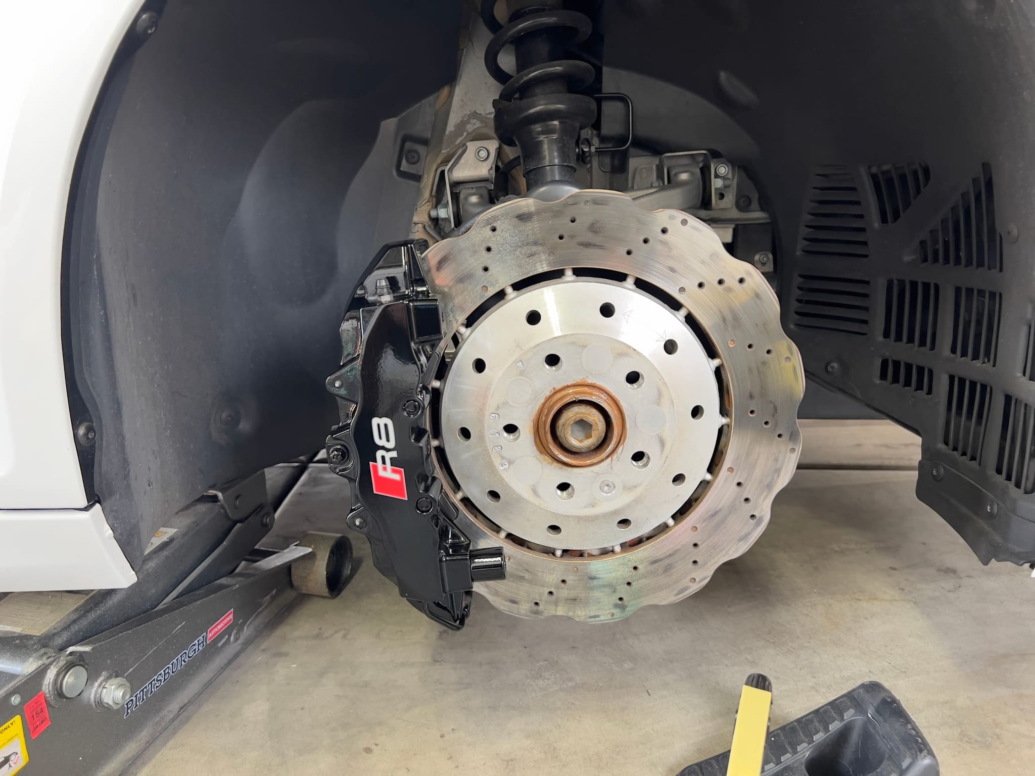 A cleaned brake rotor and caliper on a white Audi R8 with the wheel off