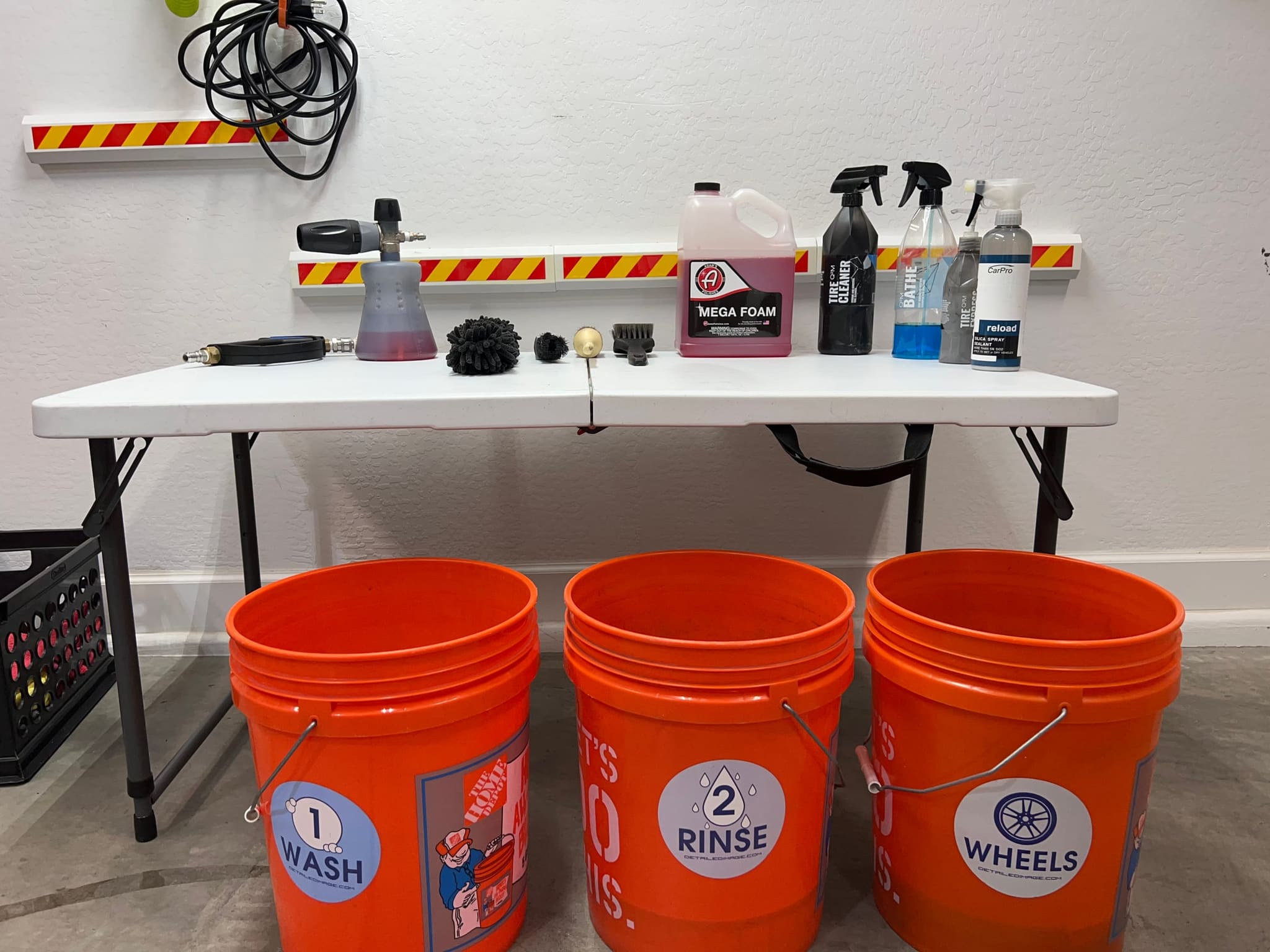 Three orange buckets labeled 1 Wash, 2 Rinse and Wheels, on a folding table of soap, brushes and a foam cannon