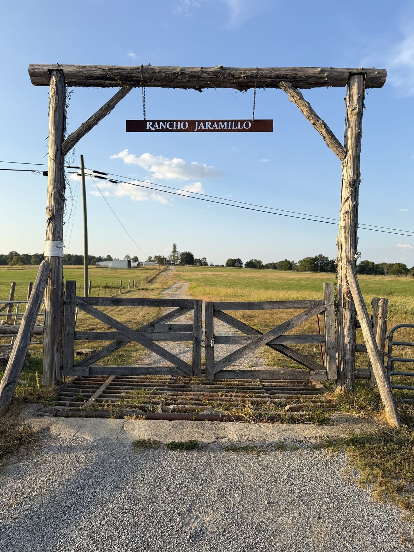 The log gate and hanging sign of Rancho Jaramillo, with a cattle guard and open pasture beyond
