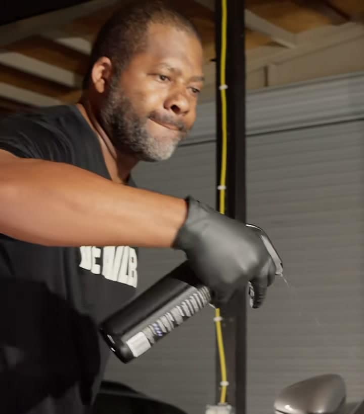 Gavin Brooks in gloves and a DETAILER shirt, misting ALL JACK onto a silver Porsche in the garage